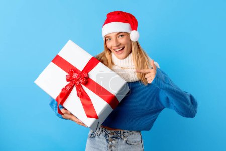 Téléchargez les photos : Heureuse dame européenne en chapeau de Père Noël tenant enveloppé boîte cadeau et pointant du doigt sur elle, debout sur fond bleu et souriant à la caméra. Concept de célébration des fêtes - en image libre de droit