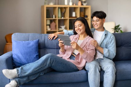 Téléchargez les photos : Heureux époux japonais, homme et femme, regardant une vidéo en ligne à l'aide d'un ordinateur tablette, partageant un temps de loisirs joyeux dans leur salon élégant. Adopter le concept de mode de vie numérique - en image libre de droit