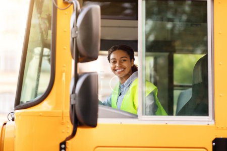 Foto de Feliz joven conductor negro sonriendo a la cámara a través de la ventana abierta mientras conduce el autobús escolar amarillo, Amistosa dama afroamericana con uniforme posando en el asiento de los conductores, disfrutando de su profesión - Imagen libre de derechos