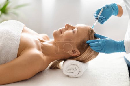 Photo for Beautiful middle aged woman receiving hair mesotherapy injection from competent beautician, attractive mature female aiming for hair rejuvenation, lying on table in modern spa salon, side view - Royalty Free Image