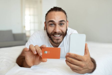 Photo for Cheerful Millennial Man Using Mobile Banking Holding Smartphone And Credit Card, Lying In Bed At Home On Weekend. Online Shopping Application, E-Commerce Concept. Selective Focus - Royalty Free Image