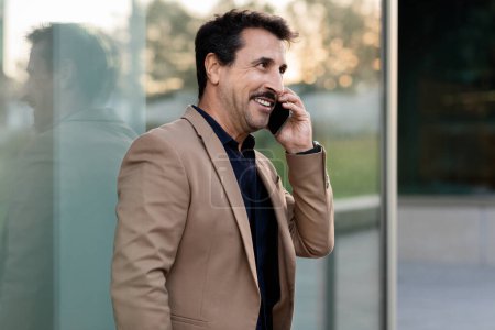 Photo for Cheerful middle-aged businessman, smoothly navigating the bustling downtown streets, is deeply engrossed in phone conversation with business partner, client, embodying the blend of dynamic - Royalty Free Image