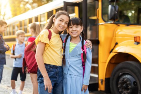 Téléchargez les photos : Enfants heureux posant près du bus scolaire jaune, prêts à rentrer à la maison après les cours, joyeux pré-adolescentes multiethniques portant des sacs à dos embrassant et souriant à la caméra, profitant de leur amitié - en image libre de droit
