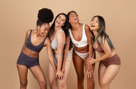 Foto de Cuatro mujeres diversas, encarnando la belleza natural, participan en poses alegres y riendo, elaborando una escena animada y auténticamente feliz en el estudio sobre fondo beige - Imagen libre de derechos