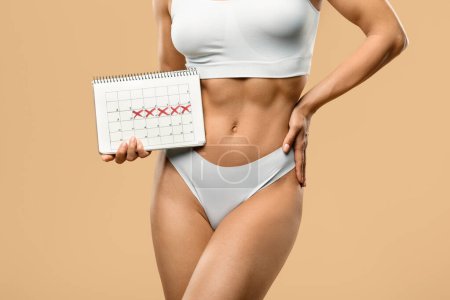 Photo for Female Cycle, Period And Menstruation Control Concept. Cropped shot of young slender lady in white underwear holding calendar with marked dates, unrecognizable woman posing over beige background - Royalty Free Image