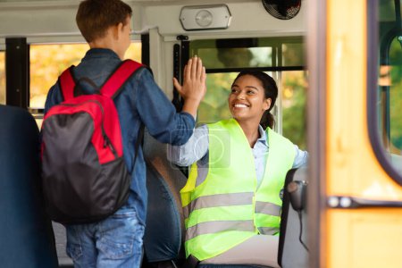 Foto de Conductora negra sonriente dando choco amistoso de cinco a niño alegre mientras entra en el autobús escolar, chófer señora en uniforme saludo alumno niño con mochila, listo para el viaje juntos - Imagen libre de derechos