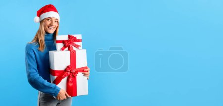 Foto de Navidad, venta de Año Nuevo. Feliz dama en Santa sombrero sosteniendo cajas de regalo y sonriendo a la cámara en el fondo del estudio azul, panorama, espacio libre. Celebración de Navidad, vacaciones de invierno humor - Imagen libre de derechos