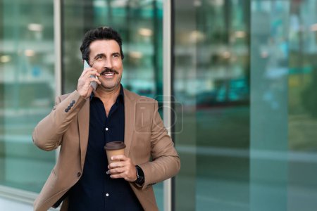Foto de El hombre de negocios de mediana edad mantiene una comunicación empresarial enfocada, participa en una conversación telefónica mientras camina por el centro de la ciudad, bebe café para llevar, mira el espacio de copia - Imagen libre de derechos