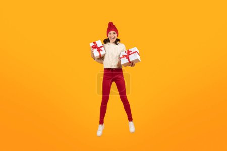 Téléchargez les photos : Joyeuse femme européenne portant un chapeau tricoté tout en sautant tenant deux boîtes cadeaux de Noël, en studio sur fond jaune. Célébrations des fêtes, saison hivernale et temps présent - en image libre de droit