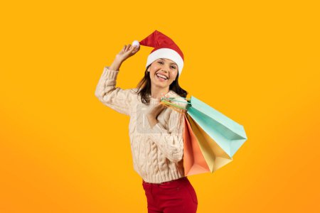 Téléchargez les photos : Joyeuse dame au chapeau de Père Noël tenant des sacs cadeaux de vacances, profitant des achats de Noël et des célébrations d'hiver sur fond de studio jaune, souriant à la caméra. Vacances d'hiver soldes et offres shopping - en image libre de droit