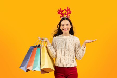 Téléchargez les photos : Shopping de Noël. Joyeuse jeune femme tenant des sacs à provisions en papier coloré achetant des cadeaux de Noël, posant sur fond de studio jaune, portant des cornes de cerf rouges drôles, souriant à la caméra - en image libre de droit