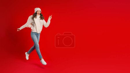 Téléchargez les photos : Super offre de Noël. Jeune femme excitée dans Santa Hat et tricot courant et sautant par-dessus le fond du studio rouge, regardant à côté de l'espace libre pour le texte, la publicité des ventes de Noël. Panorama - en image libre de droit