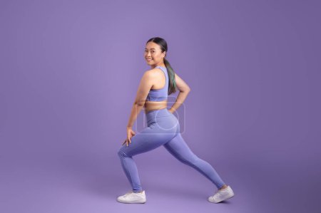 Photo for Japanese young woman in sportswear stretching legs and doing lunges over purple studio background, smiling at camera while doing exercises, full length, free space - Royalty Free Image