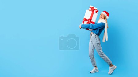 Téléchargez les photos : Jeune femme portant le chapeau de Père Noël, marchant tout en tenant pile de cadeaux de Noël, mélangeant l'esprit festif et cadeau joyeux, fond studio bleu, panorama avec espace libre - en image libre de droit