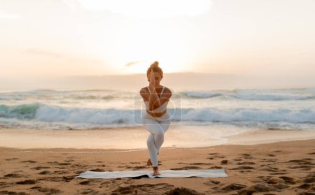 Téléchargez les photos : Femme en vêtements de sport blancs se concentre profondément comme elle tient pose de yoga, avec le coucher du soleil jetant une lueur chaude sur les vagues qui s'écrasent et le rivage sablonneux derrière elle - en image libre de droit