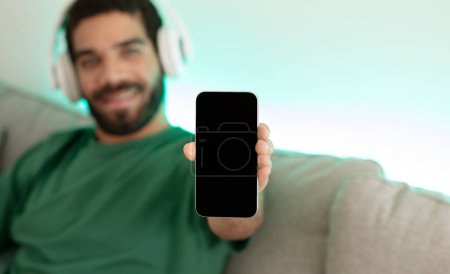 Photo for Smiling young arabic man in wireless headphones listen audio app, enjoys music, show phone with empty screen, sits on sofa, rests in living room interior. Recommendation device for lesson - Royalty Free Image