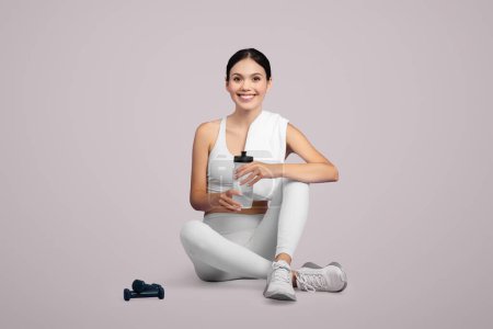Téléchargez les photos : Joyeux jeune femme en tenue de sport blanche assise les jambes croisées, tenant une bouteille d'eau, faisant une pause après l'exercice, dans un contexte de lumière apaisante, espace de copie - en image libre de droit