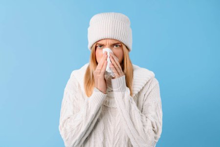 Photo for Sad angry lady in sweater and hat blowing nose in napkin on blue wall background, looking at camera. Illness, flu, cold, virus and health problems in winter - Royalty Free Image