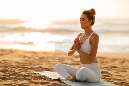 Foto de Mujer europea joven pacífica en traje blanco se sienta en meditación en la estera de yoga con los ojos cerrados, rodeado por el suave resplandor de la puesta de sol de la playa, espacio libre - Imagen libre de derechos