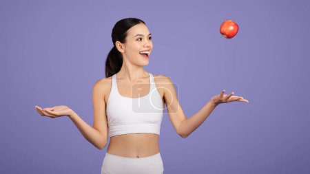 Photo for Sportive European woman in fit attire highlights a healthy lifestyle by playfully tossing red apple in studio over purple background, panorama, free space - Royalty Free Image