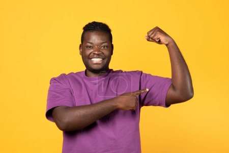 Photo for Athletic black millennial man in casual showing his muscles over yellow studio background, pointing at biceps and smiling to camera, recommending his strength workout program - Royalty Free Image