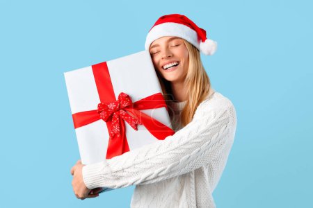 Photo for Gorgeous lady holding and hugging big gift box, posing wearing Santa hat on blue studio background. Happy woman with Christmas present ready to celebrate Xmas or New Year - Royalty Free Image