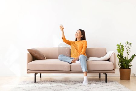 Photo for Happy pretty young asian woman wearing comfy casual outfit sitting on couch at home, holding remote control and looking at empty space over white empty wall, turning on AC - Royalty Free Image