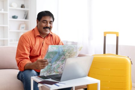 Photo for Travel Destination Choice. Excited Mature Indian Man Tourist Looking At Paper Map Planning His Route, Sitting With Laptop And Suitcase Indoor At Modern Home Interior, Copy Space - Royalty Free Image