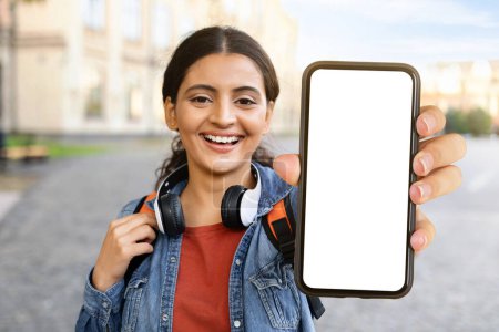 Photo for Young indian woman student showing smartphone with white blank screen, posing at campus, copy space, mockup. Positive cheerful lady with backpack and wireless headset recommending educational app - Royalty Free Image