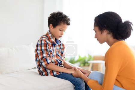 Photo for Mommy comforting distressed son boy indoors, holding hands in deep conversation, addressing challenges like school bullying and family disputes. Side view. Parental support - Royalty Free Image