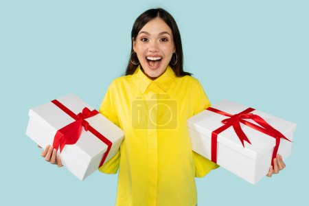 Photo for Emotional young brunette woman wearing basic yellow shirt standing hold white present boxes with red gift ribbon bow looking at camera isolated on blue colour background, birthday celebration - Royalty Free Image