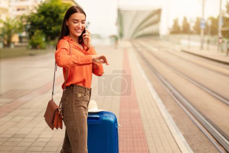 Photo for Lady glancing at her watch talking on phone, anticipating tram arrival in city, standing solo with suitcase luggage at a streetcar stop in urban area. Commuting and transport time schedules - Royalty Free Image