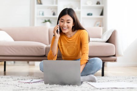 Photo for Cheerful busy young asian woman freelancer working from home. Happy smiling korean lady sitting on floor in living room, have phone conversation with client, looking at laptop screen, copy space - Royalty Free Image