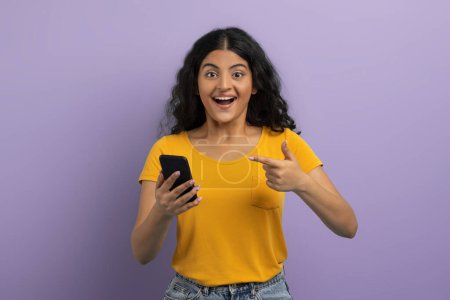 Téléchargez les photos : Joyeux joyeux joyeux jeune étudiante indienne bouclée pointant le téléphone dans sa main et grimaçant, vérifier belle offre en ligne, affaire, fond de studio violet. Application mobile - en image libre de droit