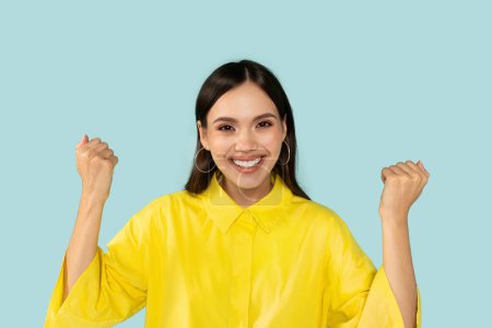 Photo for Emotional happy beautiful young brunette lady wearing yellow shirt celebrating success, great news, clenching fists and smiling at camera isolated on blue studio background - Royalty Free Image