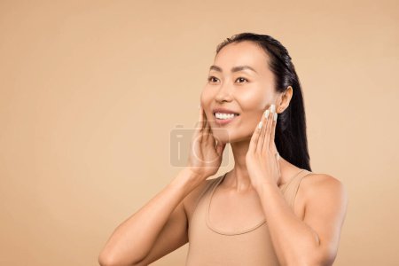 Téléchargez les photos : Femme asiatique souriante à la peau rayonnante, touche son visage, illustrant les résultats des soins de la peau et la beauté naturelle sur fond beige, espace libre - en image libre de droit
