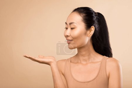 Foto de Mujer asiática serena de mediana edad, extendiendo su mano y presentando un nuevo producto de belleza en su palma, filmada en un estudio con un fondo beige suave - Imagen libre de derechos