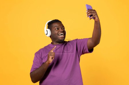 Photo for Telecommunication concept. Cheerful young black guy in purple t shirt using brand new wireless headset and smartphone on yellow studio background, having video call with friends - Royalty Free Image