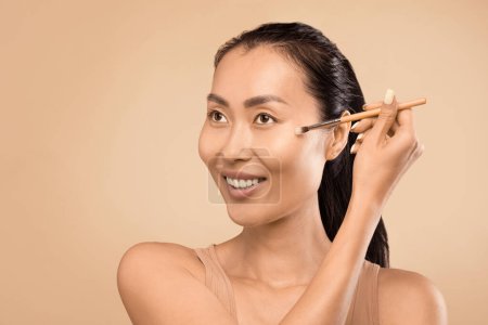 Téléchargez les photos : Gracieuse femme asiatique d'âge moyen perfectionne sa routine beauté, brossant doucement sur surligneur dans le cadre de son application de maquillage nu, soulignant son élégance naturelle - en image libre de droit