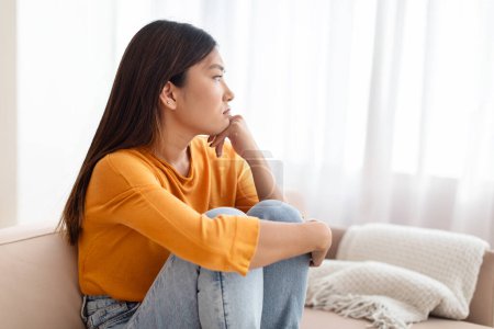 Téléchargez les photos : Malheureuse femme asiatique millénaire assise seule sur le canapé à la maison, embrassant ses genoux, regardant par la fenêtre, souffrant de dépression, anxiété, dépression mentale, espace de copie - en image libre de droit