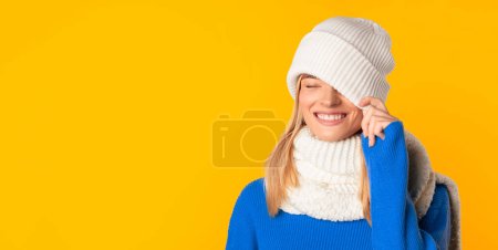 Photo for Bright winter offer. Carefree happy lady covering one eye with white knitted hat, posing over yellow studio background, panorama with copy space - Royalty Free Image