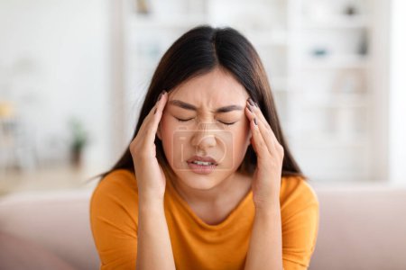 Photo for Unhappy long-haired young asian lady sitting on couch at home, rubbing her temples, closed eyes, suffering from headache, chinese woman experiencing difficulties, copy space, closeup - Royalty Free Image