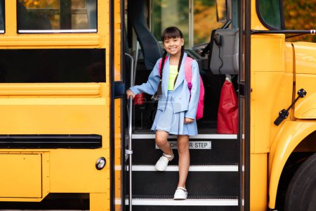 Foto de Sonriente niña asiática preadolescente bajándose del autobús escolar amarillo, niña feliz con mochila bajando del vehículo mientras llega para estudiar, lista para las lecciones y aprender diversión - Imagen libre de derechos