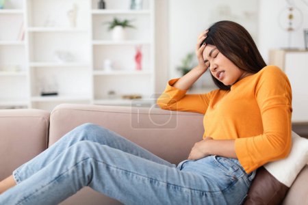 Foto de Enfermedad de Gastritis. Joven mujer china que sufre de dolor de estómago, tocándose el estómago mientras tiene un espasmo doloroso en el vientre sentado en el sofá en casa. Síntoma de dolor abdominal, concepto de problemas de salud - Imagen libre de derechos