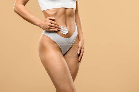 Photo for Unrecognizable female in white underwear applying moisturizing body lotion on belly, slim woman with sporty figure rubbing fat burning cream on tummy while standing on beige background, copy space - Royalty Free Image