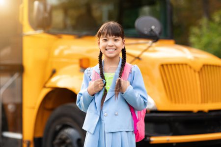 Foto de Retrato de feliz sonrisa chica asiática posando además de autobús escolar amarillo al aire libre, alegre lindo niño femenino con mochila mirando a la cámara, listo para las clases y aprender diversión, espacio de copia - Imagen libre de derechos