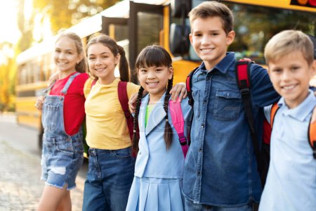 Foto de Niños alegres listos para abordar el autobús escolar, grupo de alumnos felices con mochilas al aire libre, abrazando y sonriendo a la cámara, posando cerca del autobús escolar después de las lecciones, prepararse para el viaje a casa - Imagen libre de derechos