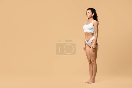 Photo for Young slender woman with perfect body shape posing in white underwear over beige studio background, smiling female reveal sculpted figure, looking aside at copy space, full length shot - Royalty Free Image