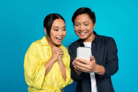 Téléchargez les photos : Surpris inspiré couple asiatique gai dans casual regardant smartphone, se réjouissant de bonnes nouvelles, isolé sur fond de studio bleu. App pour les achats en ligne, la publicité et l'offre, discuter dans les réseaux sociaux - en image libre de droit