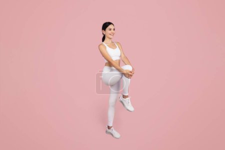 Téléchargez les photos : Femme en forme et athlétique en vêtements de sport blancs étirant sa jambe et se réchauffant, sur fond rose doux, mettant en valeur la santé et la flexibilité - en image libre de droit